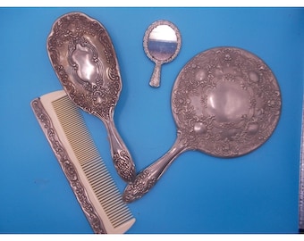 May include: A vanity set consisting of an oval hand mirror, a smaller round hand mirror, and a rectangular comb. The mirrors feature ornate silver-toned metal backs with intricate embossed floral patterns, while the comb has a matching silver-toned handle.