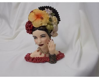 May include: A decorative figurine of a person's head and shoulders, featuring dark hair adorned with large yellow, red, and white flowers. The figure wears a red ruffled off-the-shoulder top, a beaded necklace, a dark bracelet, and has red nail polish.