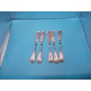 May include: A set of five silver-toned butter knives arranged in a row. Each knife features a smooth, rounded blade and a handle decorated with a subtle, embossed shell motif at the base. The metal has a polished finish with visible vertical lines.