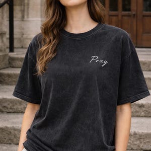Op de afbeelding: Donkergrijs T-shirt met korte mouwen. Het woord "Pray" is in witte letters op de bovenborst geborduurd. Het shirt heeft een licht vervaagde, vintage look.