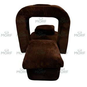 K&ouml;nnte beinhalten: Ein brauner Cord-Sessel mit einem einzigartigen, geschwungenen R&uuml;cken und Armlehnen. Der Stuhl hat eine rechteckige Sitzfl&auml;che und eine separate Kopfst&uuml;tze. Der Stoff hat eine vertikale Rippenstruktur. Das Wort "MORF" ist auf dem Stuhl aufgedruckt.