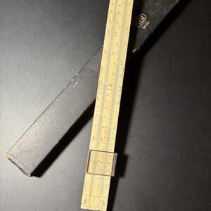 Peut inclure: Une règle à calcul beige avec un curseur en plastique transparent et des marquages numériques noirs, accompagnée d'un étui de protection noir texturé. La règle porte les inscriptions "HEMMI" et "1448" sur l'étui, et "MADE IN JAPAN" sur la règle.