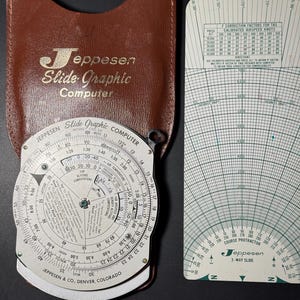 May include: A Jeppesen Slide Graphic Computer with a brown leather case and a separate white plastic 3-way slide course protractor. The circular computer features black text, numerical scales, and a central rotating dial for aviation calculations.