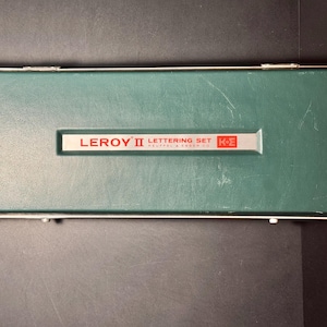 May include: A rectangular green storage case with a silver metal frame and two hinges. A central silver label features the red text LEROY II LETTERING SET KEUFFEL & ESSER CO and a red square logo with the letters K and E inside.