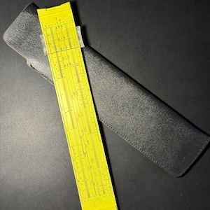 May include: A yellow plastic slide rule featuring multiple numerical scales and markings, positioned diagonally across a black textured protective case. The slide rule displays various mathematical notations and precise black printed graduations.