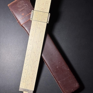 May include: A beige slide rule with a clear plastic cursor and metal end caps, featuring printed numerical scales and the brand name POST. It is accompanied by a dark brown leather protective case with a flap and embossed logo.