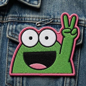 May include: Embroidered patch featuring a green frog with white eyes, a black mouth, and a pink outline. The frog is giving a peace sign with its right hand. A silver safety pin is pinned above the patch on a denim jacket.