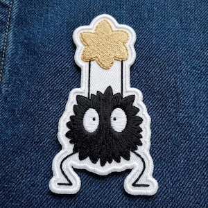 May include: Embroidered patch featuring a black, round, fuzzy creature with large white eyes and a gold star-shaped object above it. The patch has a white border and is on a blue denim background.