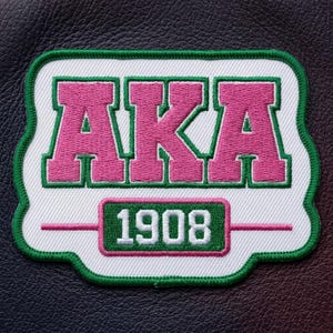 May include: Embroidered patch featuring the letters "AKA" in pink with green trim, set against a white background. The year "1908" is displayed below in a green rectangle. The patch has a green border and is set on a dark leather-like surface.