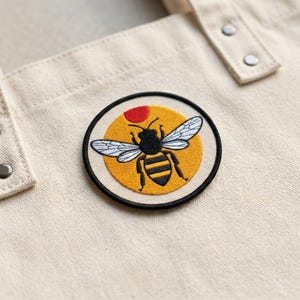 May include: A round embroidered patch featuring a stylized bee with white wings and black and yellow stripes. The bee is set against a yellow background with a red dot, all within a black border. The patch is attached to a beige tote bag.