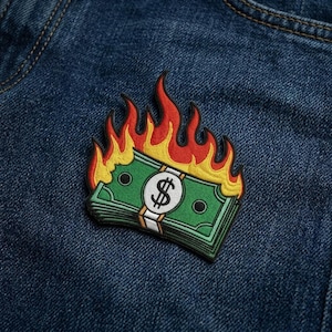 May include: Embroidered patch featuring a stack of green dollar bills with a white band and a dollar sign, engulfed in flames. The flames are red and yellow, creating a bold visual on a blue denim background.