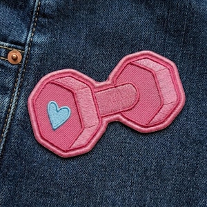 May include: Pink embroidered patch in the shape of a dumbbell with a light blue heart on one end. The patch is on a dark blue denim background. The dumbbell design has a geometric shape.