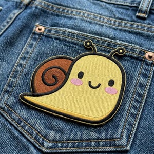 May include: A yellow embroidered snail patch with a brown shell, black outlines, and pink cheeks is affixed to the back pocket of a pair of blue denim jeans. The snail has a smiling face and is a cute, whimsical design.