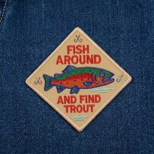 May include: A diamond-shaped patch with the text "FISH AROUND AND FIND TROUT" and a colorful fish illustration. The patch is tan with red, green, blue, and orange details, and is set against a blue denim background.