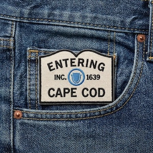 May include: Close-up of a blue denim pocket with a rectangular patch. The patch is white with black text that reads "ENTERING INC. 1639 CAPE COD." A blue emblem is in the center. The pocket has yellow stitching and a copper rivet.