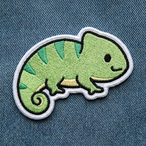 May include: Embroidered patch featuring a cartoon chameleon in shades of green with a white border. The chameleon has a smiling face and a curled tail. The patch is on a blue denim background.