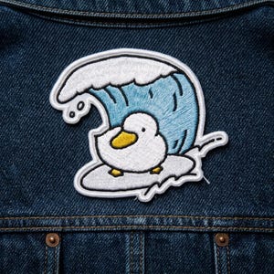 May include: Embroidered patch featuring a cartoon duck surfing a wave. The duck is white with a yellow beak and feet, and the wave is blue and white. The patch is on a blue denim background.