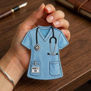 May include: A light blue embroidered patch in the shape of a medical scrub top, featuring a black stethoscope and a pocket with the name "NANCY, RN". The patch is held in a hand.
