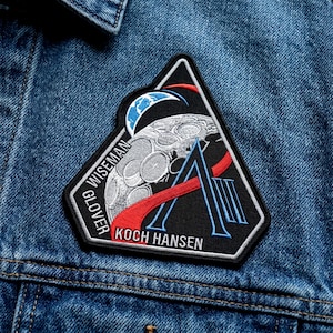 May include: A black, triangular embroidered patch featuring a stylized moon, red and blue accents, and the names "WISEMAN", "GLOVER", "KOCH HANSEN". The patch is sewn onto a denim jacket.