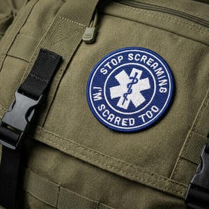May include: A round, blue and white embroidered patch with the text "STOP SCREAMING I'M SCARED TOO" encircling a white medical symbol. The patch is affixed to an olive green tactical backpack.