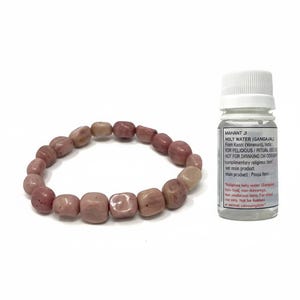 May include: A pink and beige beaded bracelet next to a small bottle with a white cap. The bottle has a label with black text. The bracelet is made of square-shaped beads.