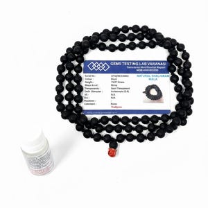 May include: A black mala necklace with round beads, a red accent bead, and a small white bottle. A card with text about the necklace is also present. The necklace is made of black beads.