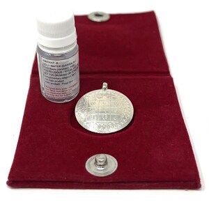 May include: A silver pendant with etched designs and a small bottle of liquid, labeled "Holy Water," rest inside a red velvet pouch. The bottle has a white cap and text. The pouch is secured with a silver snap.