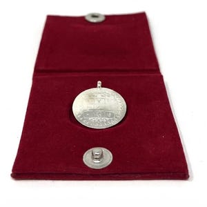 May include: A silver pendant with intricate designs rests inside an open, burgundy velvet case. The pendant is circular with detailed engravings. The case has a magnetic closure and a silver button clasp. The background is white.