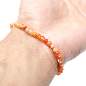May include: A delicate bracelet composed of small, round beads in shades of orange, red, and white. The bracelet is worn on a wrist, showcasing its vibrant colors and simple design. This beaded jewelry is a stylish accessory.
