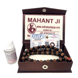 May include: A brown box with a beaded necklace inside, accompanied by a small bottle. The box has the text "MAHANT JI" and a website address. A card with text is also visible. The necklace beads are dark brown and black.