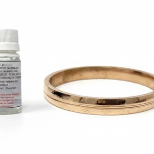 May include: A gold-colored bangle bracelet and a small bottle of holy water. The bottle has a white cap and a label with text. The bangle is a simple, round design.