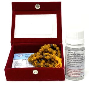 May include: A maroon velvet box with a clear lid contains a beaded necklace with yellow beads. A small bottle of holy water is next to the box. The bottle has a white cap and a label with text.