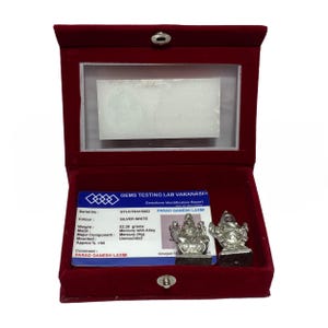 May include: A silver-colored figurine of a deity, displayed in a red velvet box with a clear lid. The box contains a gemstone identification report and a second figurine. The figurines are on a dark base.