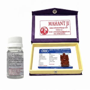 May include: A clear bottle of holy water labeled "MAHANT JI HOLY WATER (GANGAJAL)" sits next to an open purple box. Inside the box is a small brown statue of a deity and a card with text. The box also has the text "MAHANT JI" and contact information.