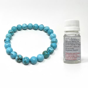 May include: A turquoise beaded bracelet and a small white bottle with text. The bracelet is made of round, light blue beads with dark blue and black marbling. The bottle has a white cap and a label with black text.
