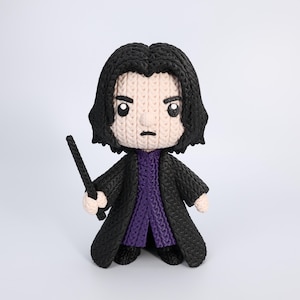 May include: A handmade, knitted doll of a character with black hair, wearing a black coat over a purple sweater, and holding a wand. The doll has a light skin tone and is set against a white background.