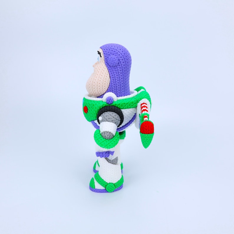 May include: A crocheted figurine of a space ranger character featuring a purple hood, a beige face, and a white suit with green, red, and black accents. The figure includes a backpack with red and white striped wings and a green base.