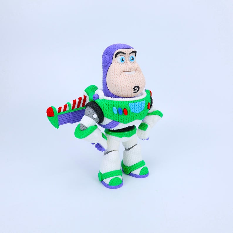 May include: A crocheted Buzz Lightyear figure featuring a purple hood, a white and green space suit with red and blue accents, and a detachable wing assembly with red and white stripes. The character has a tan face with blue eyes and a small goatee.