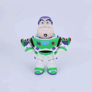 May include: A crocheted plush figure of a space ranger character with a purple hood, a white face, and a green and white chest plate. The figure features blue eyes, a small chin goatee, and purple wings extending from the back of the suit.