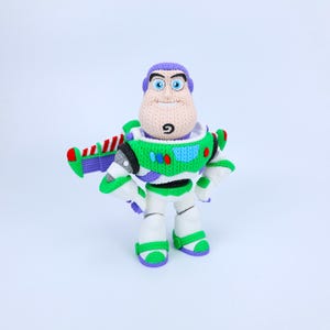 May include: A crocheted Buzz Lightyear figure featuring a purple hood, a white and green chest plate with red and blue accents, and white legs with green trim. The character has blue eyes, a tan face, and a small black goatee on the chin.
