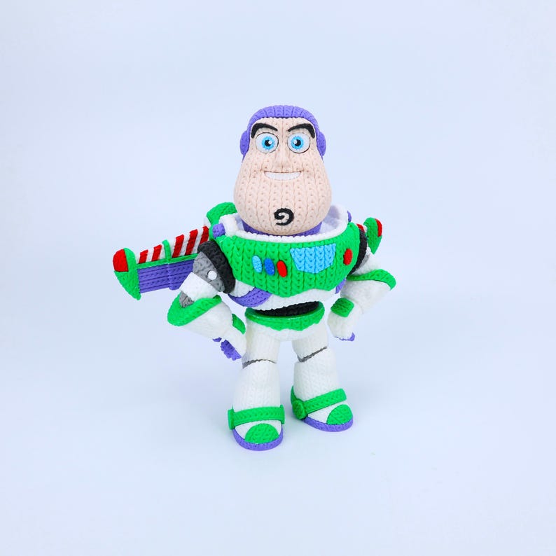 May include: A crocheted Buzz Lightyear figure featuring a purple hood, a white and green chest plate with red and blue accents, and white legs with green trim. The character has blue eyes, a tan face, and a small black goatee on the chin.