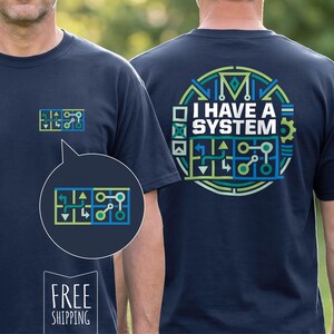 以下が含まれることがあります： ネイビーブルーのTシャツ。前面には小さくカラフルなグラフィック、背面には大きな円形デザインが施されています。背面のデザインには「I HAVE A SYSTEM」の文字と、緑、青、黄色の幾何学模様が含まれています。前面のグラフィックは、背面のデザインの縮小版です。