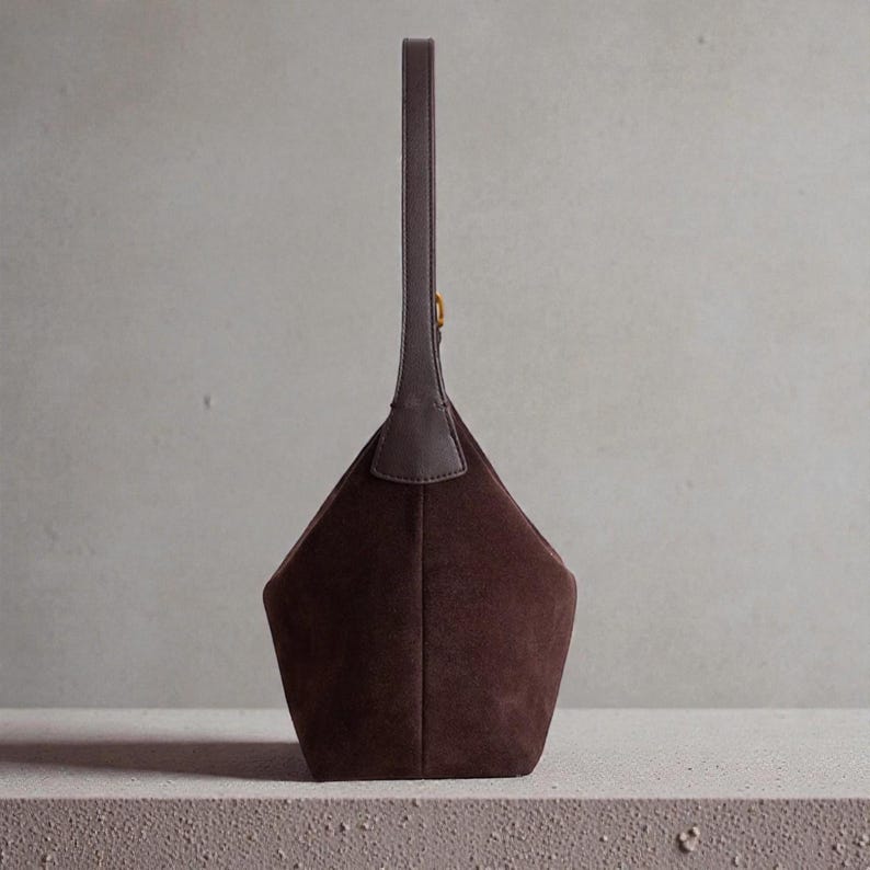 May include: A dark brown suede handbag with a geometric design. The bag has a long, slender handle and a small gold-colored accent. The bag is standing upright on a light-colored surface.