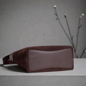 May include: A brown suede handbag with a rectangular base and a curved leather panel. The bag has a single strap and is set against a neutral background. The bag's dimensions are not visible.