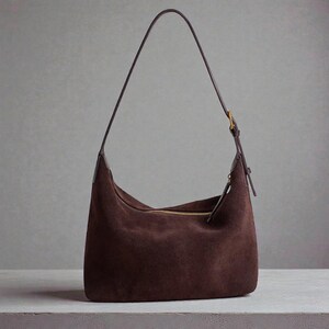 May include: A brown suede shoulder bag with a long, thin strap. The bag has a curved shape and a gold-colored zipper. The strap is attached with a gold-colored buckle.