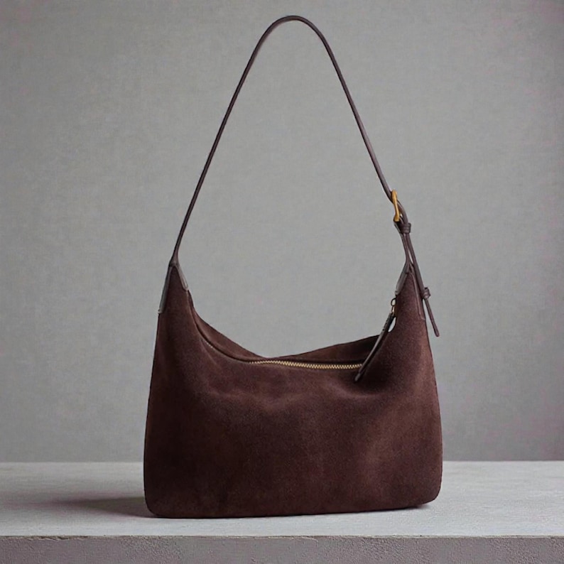 May include: A brown suede shoulder bag with a long, thin strap. The bag has a curved shape and a gold-colored zipper. The strap is attached with a gold-colored buckle.