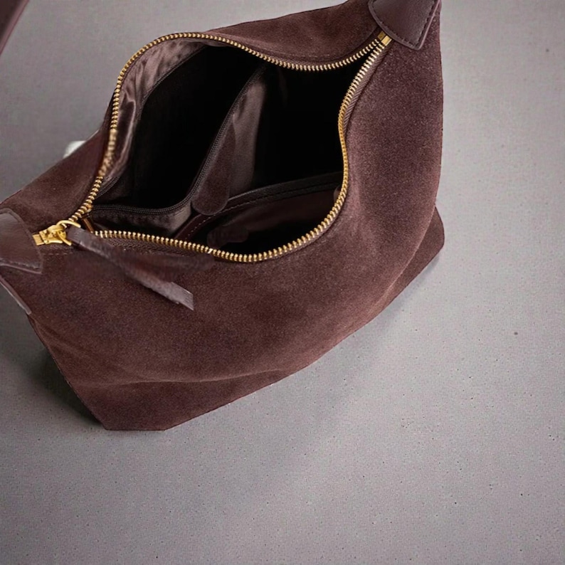 May include: A brown suede handbag with a gold zipper, open to reveal the interior compartments. The bag has a rounded shape and a dark brown leather accent at the zipper pull. The interior lining is a darker shade of brown.