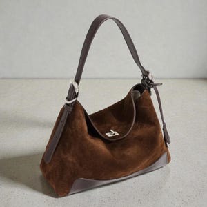Peut inclure: Un sac à bandoulière en daim marron avec une bandoulière en cuir marron foncé et des accessoires argentés. Le sac a une forme arrondie avec une fermeture à rabat et une base en cuir grise. Les dimensions du sac ne sont pas visibles.