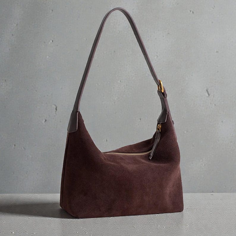 May include: A dark brown suede shoulder bag with a curved shape and a long strap. The bag has a zipper closure with a gold-colored zipper and hardware. The strap is a darker shade of brown.