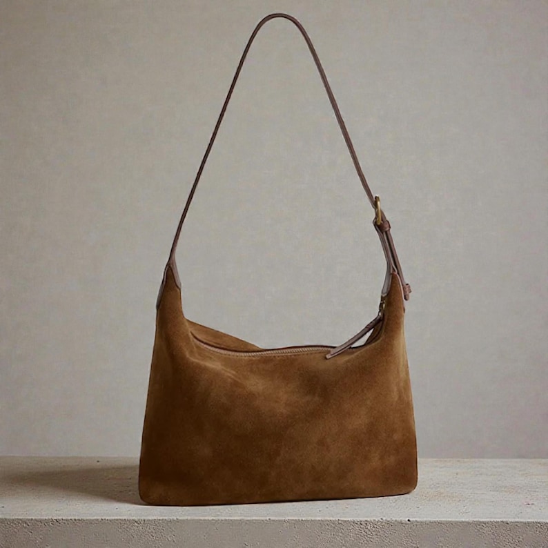 May include: A brown suede shoulder bag with a long, thin strap. The bag has a curved shape and a zip closure. The strap is a darker brown leather, and the bag appears to be sitting on a white surface.